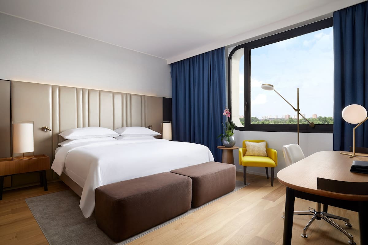 A modern hotel room with a large bed, a desk, a chair, and a view of the city through a large window.