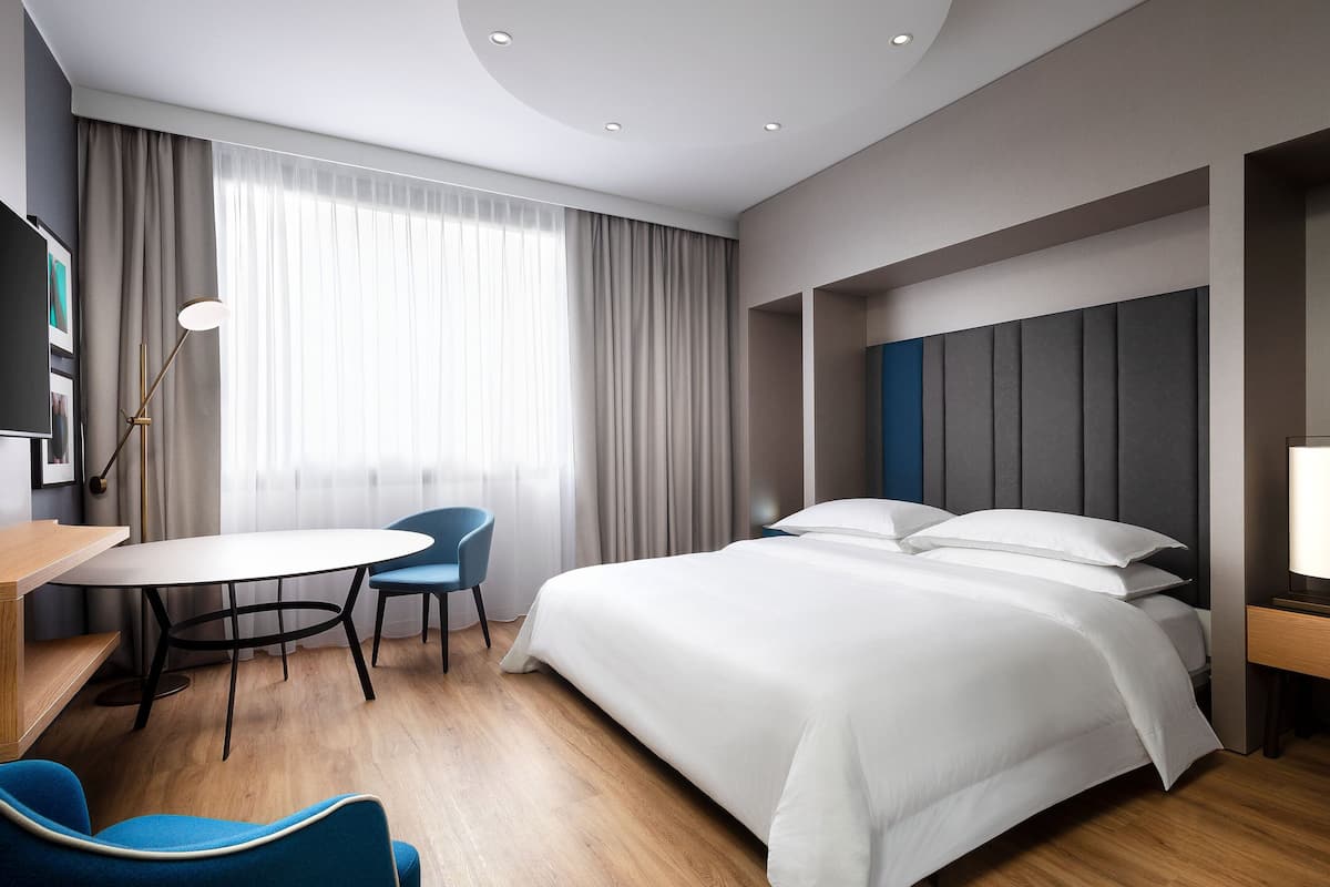 A modern hotel room with a large bed, a desk with a chair, and a large window with curtains.