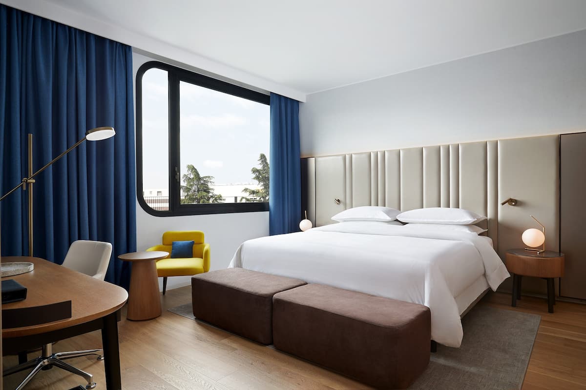 A modern hotel room with a large bed, a desk, a chair, and a view of the city.