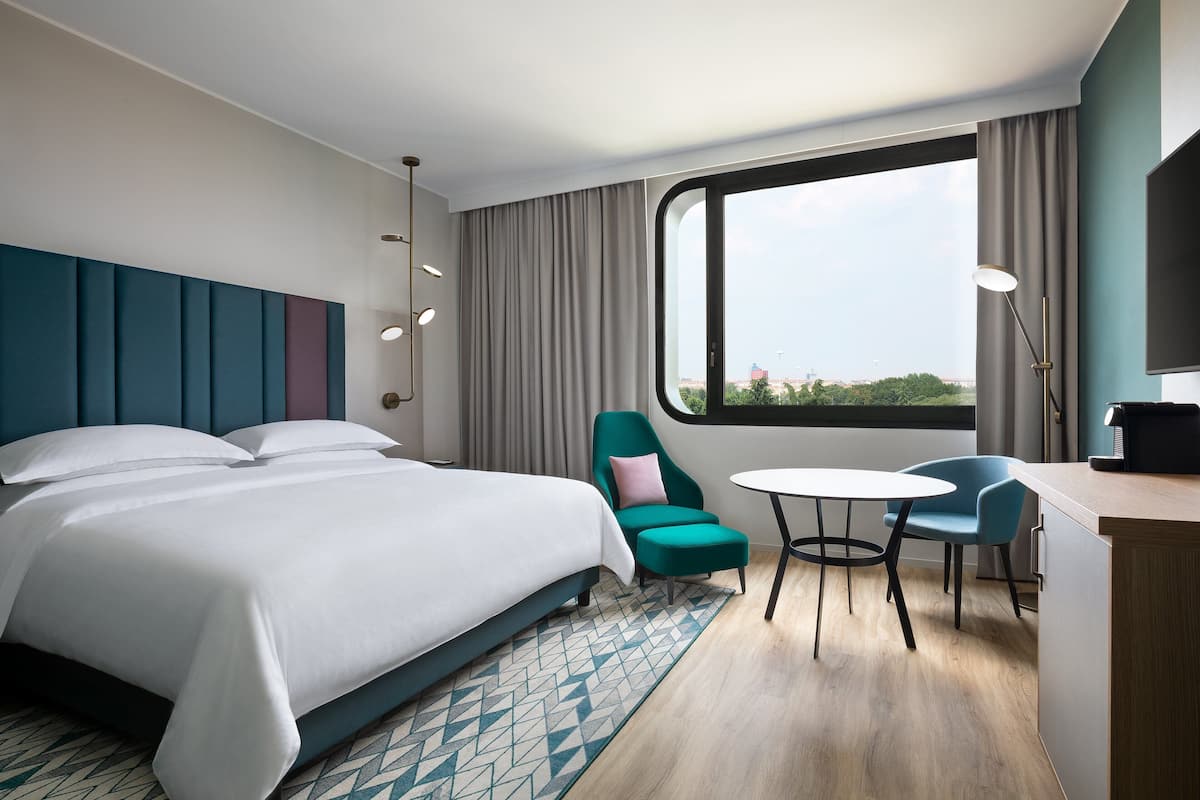 A modern hotel room with a large bed, a desk with a chair, a small table, and a view through a window.