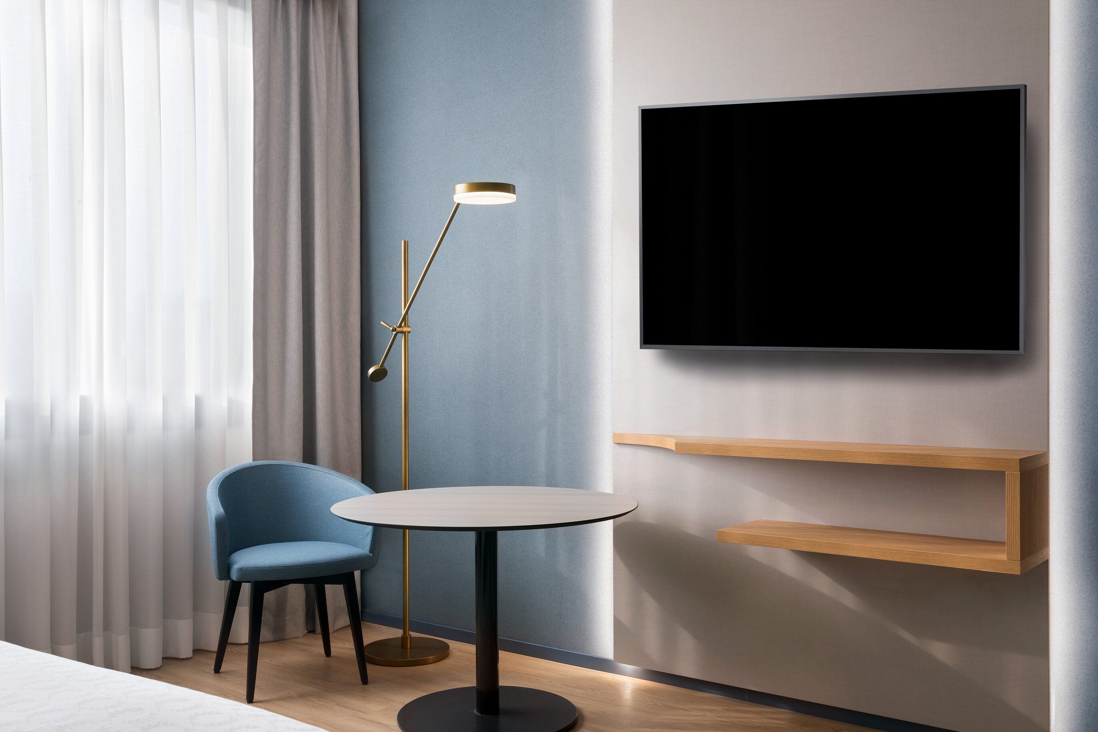 Una habitación de hotel moderna con una mesa redonda, una silla azul, una lámpara de pie, un televisor montado en la pared y estanterías de madera.