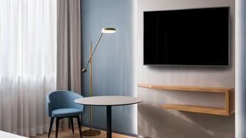 Una habitación de hotel moderna con una mesa redonda, una silla azul, una lámpara de pie, un televisor montado en la pared y estanterías de madera.