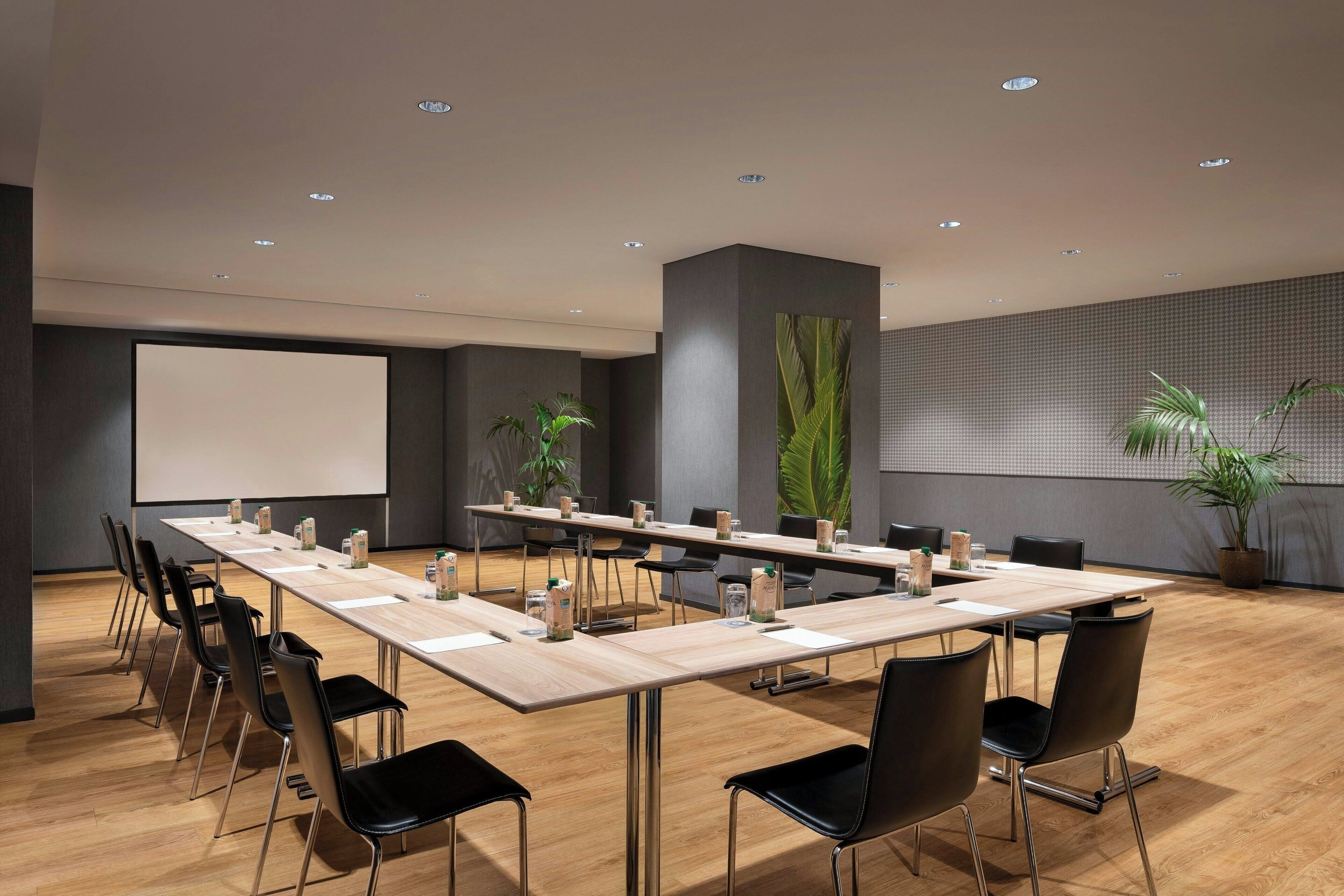 A conference room with a long table, black chairs, a projector screen, and potted plants.