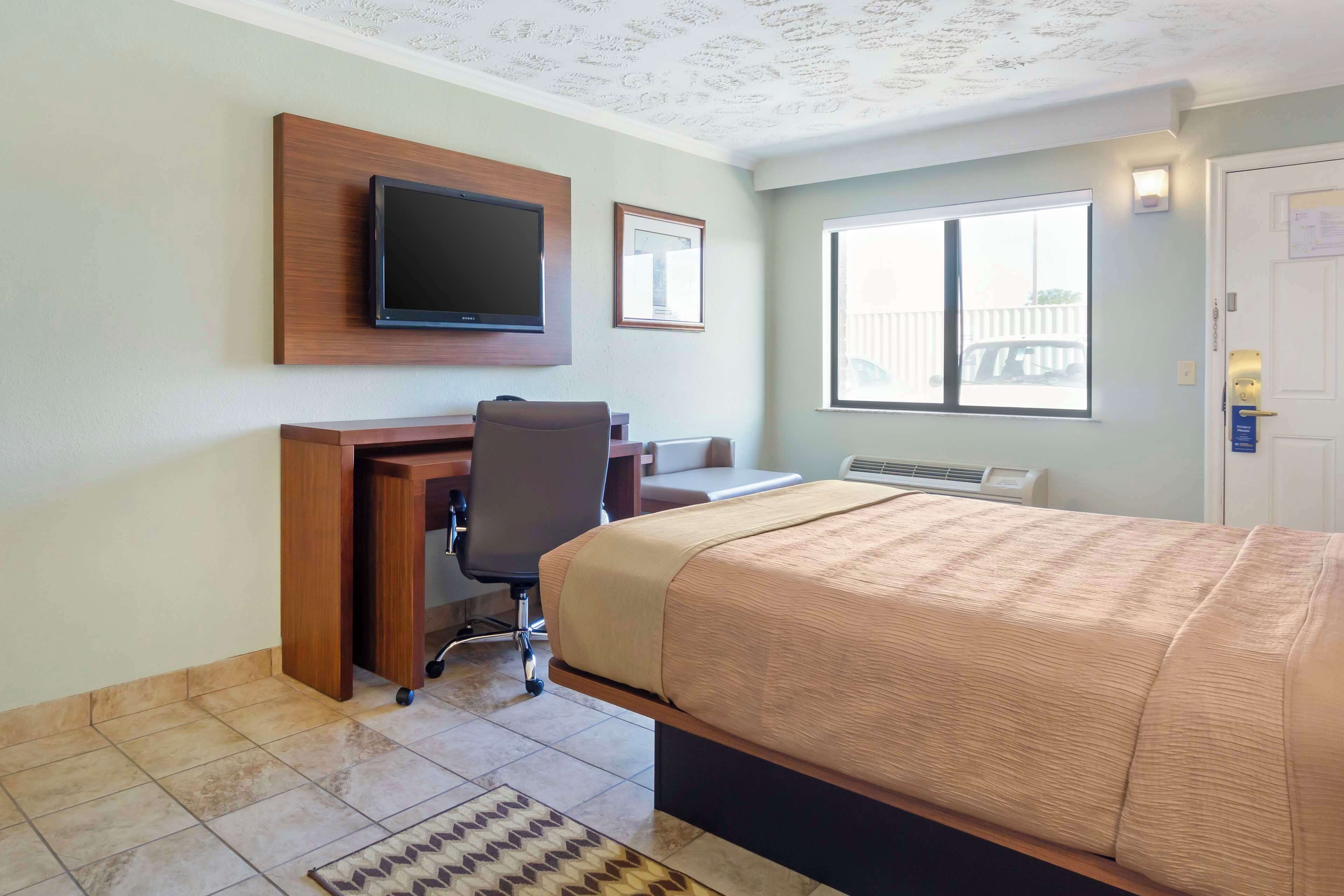 A hotel room with a bed, desk, chair, television, and a window with a view of a fence.