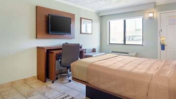 A hotel room with a bed, desk, chair, television, and a window with a view of a fence.