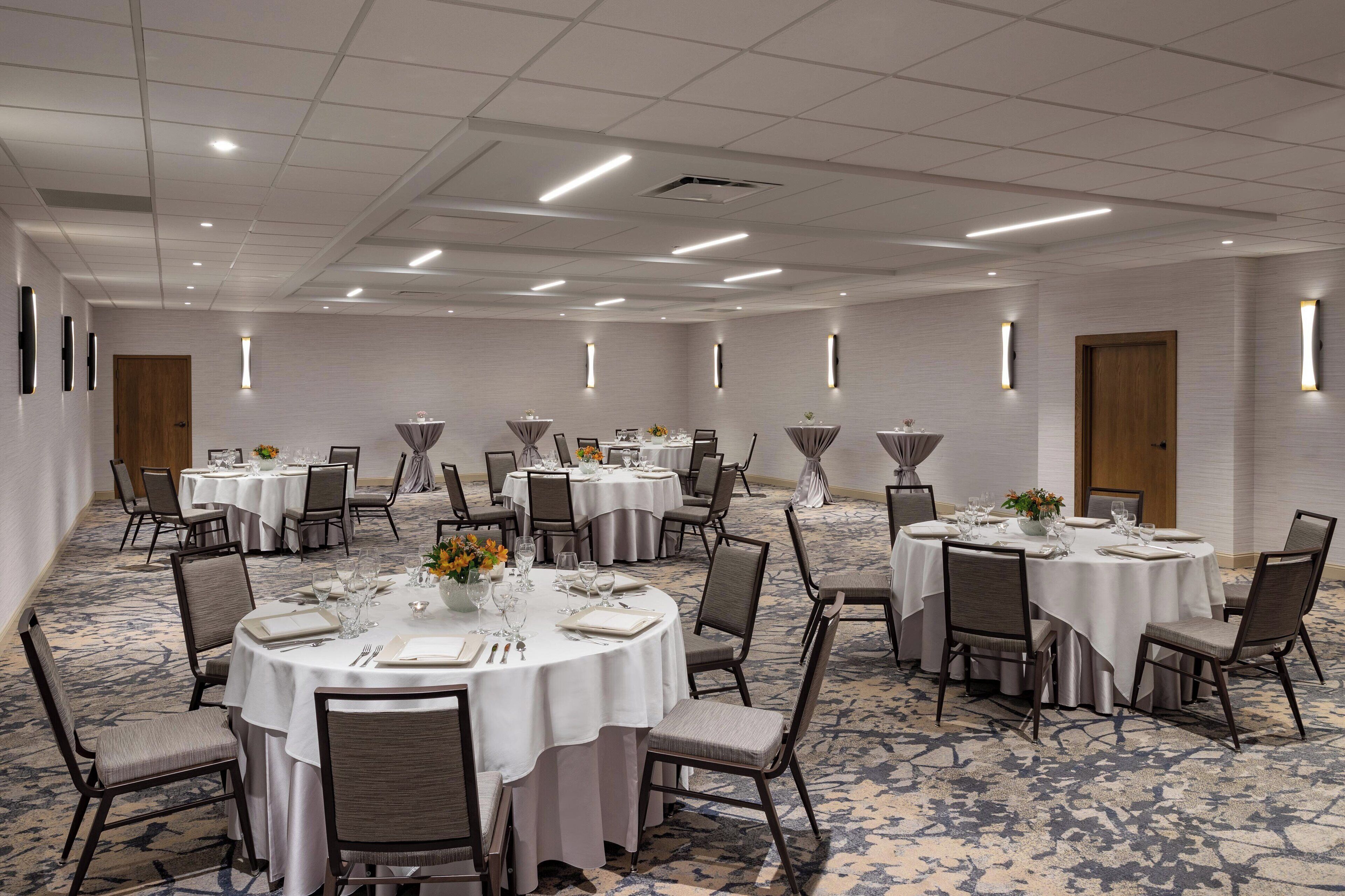 A large conference room with multiple round tables, each set with chairs, white tablecloths, and centerpieces.