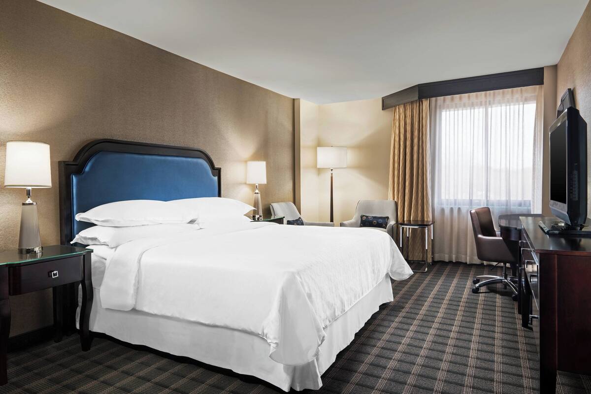 Een hotelkamer met een groot bed, twee nachtkastjes met lampen, een bureau met een stoel, een televisie en een raam met gordijnen.