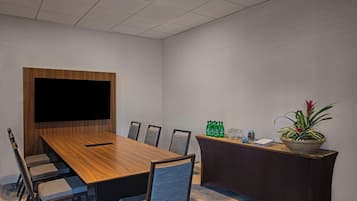 A conference room with a long wooden table, six chairs, a large flat-screen TV, a reception desk with a potted plant, and a small table with bottled water and glasses.