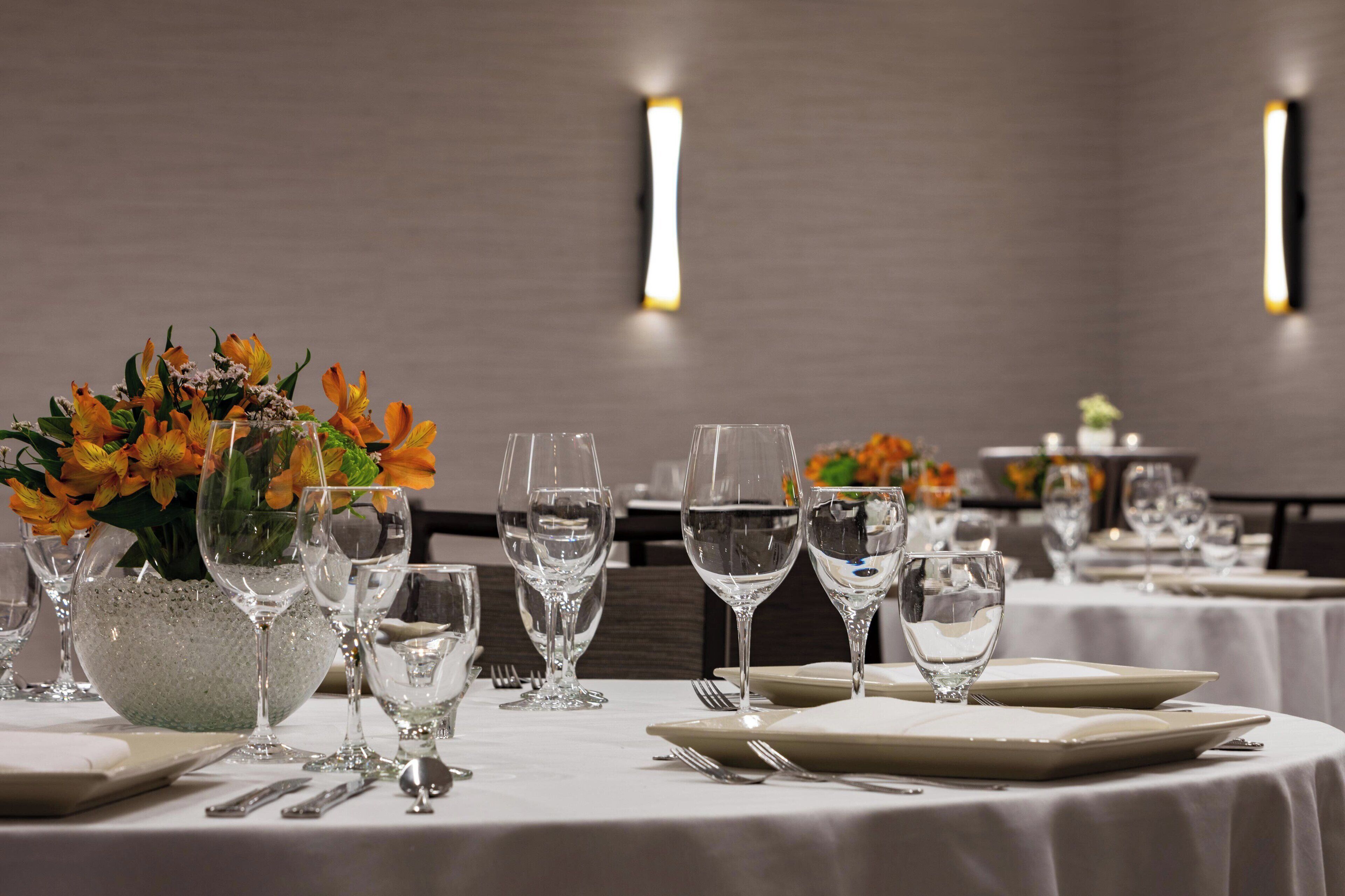 A dining table set with white tablecloths, clear glassware, and a centerpiece of orange flowers.