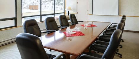 Un salón de conferencias con una mesa de madera, sillas negras, una pizarra blanca y ventanas amplias con vistas a árboles y un edificio.