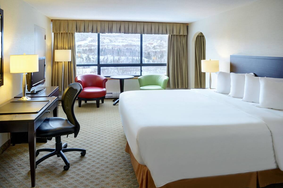Une chambre d’hôtel avec un grand lit, un bureau avec un ordinateur, une télévision, deux chaises et une fenêtre donnant sur un paysage enneigé.