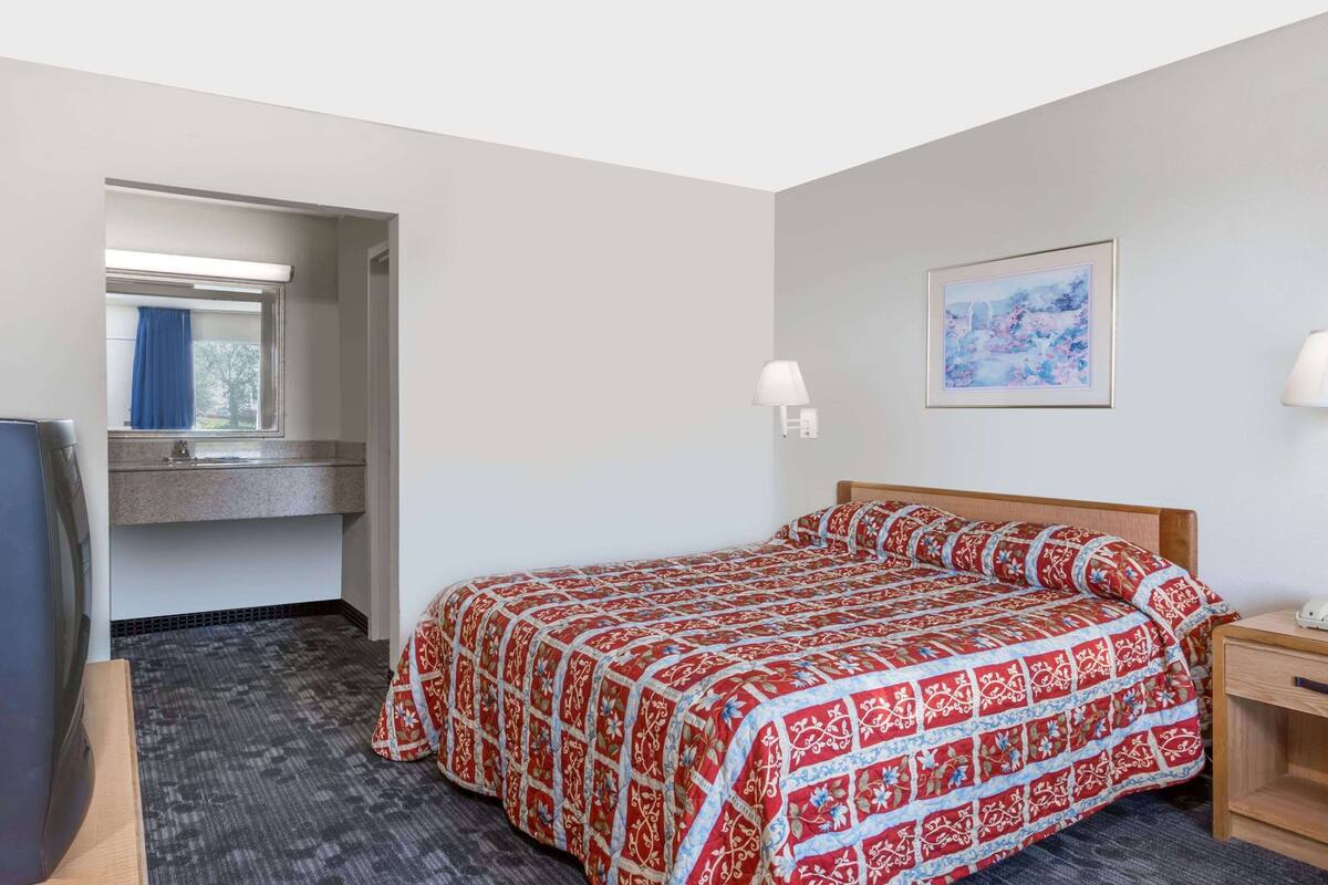 A hotel room with a bed, a desk, a chair, a window with a curtain, a wall-mounted lamp, and a painting on the wall.