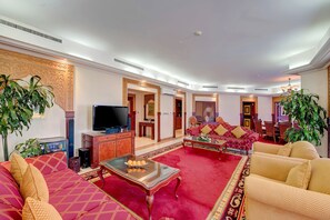 Un salón comedor amplio con alfombra roja, un conjunto de sofás, una mesa de centro con objetos decorativos y un televisor.