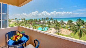 Un balcon avec une table sur laquelle sont disposés des fruits, donnant sur un espace piscine agrémenté de palmiers et d’une plage.
