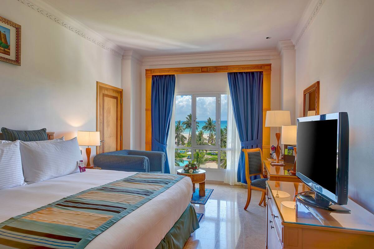 Una habitación de hotel con una cama grande, televisor, escritorio con lámpara y vista al océano a través de la ventana.