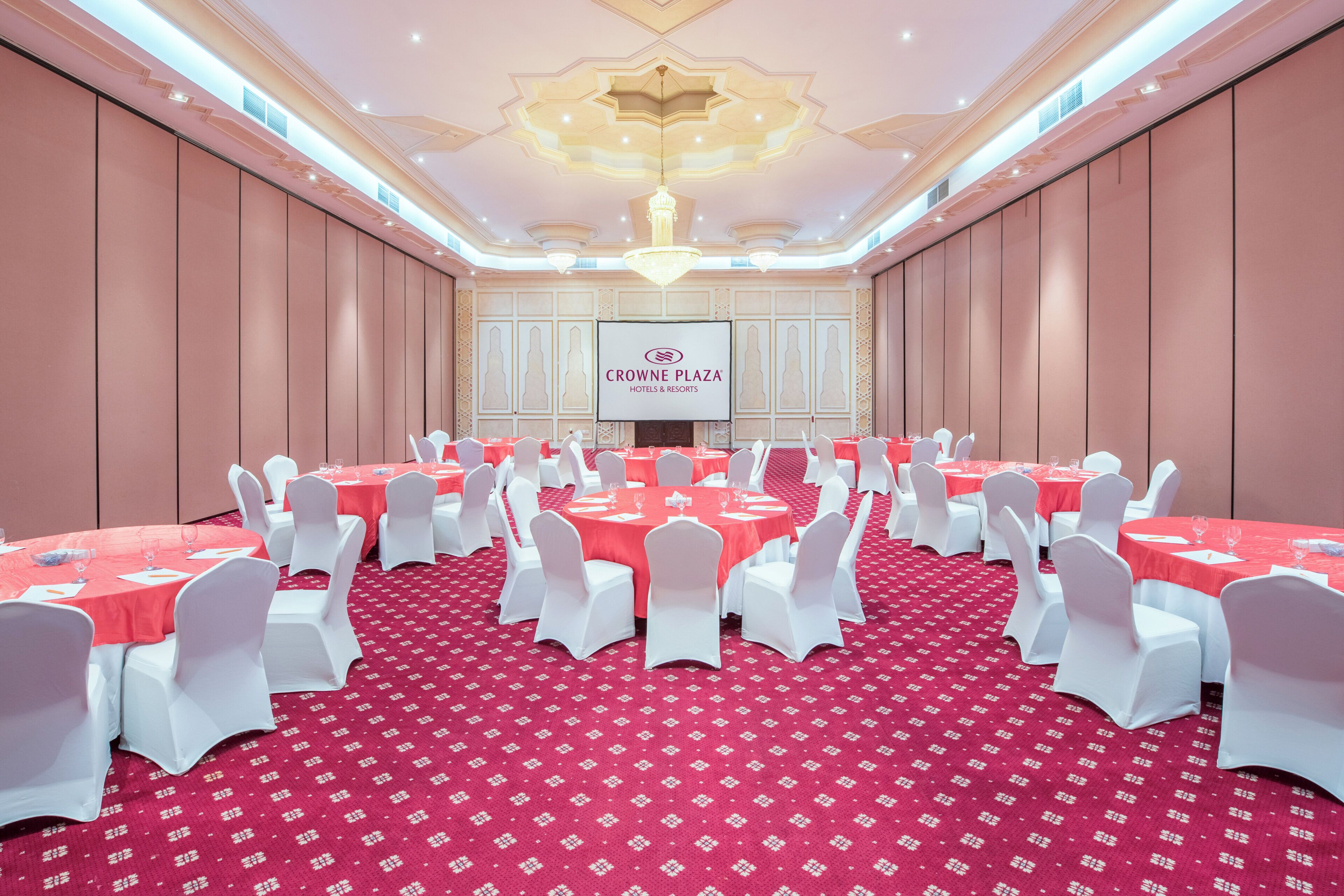 A conference room with tables set for a meeting, white chairs, a red carpet, and a large screen displaying the hotel name.