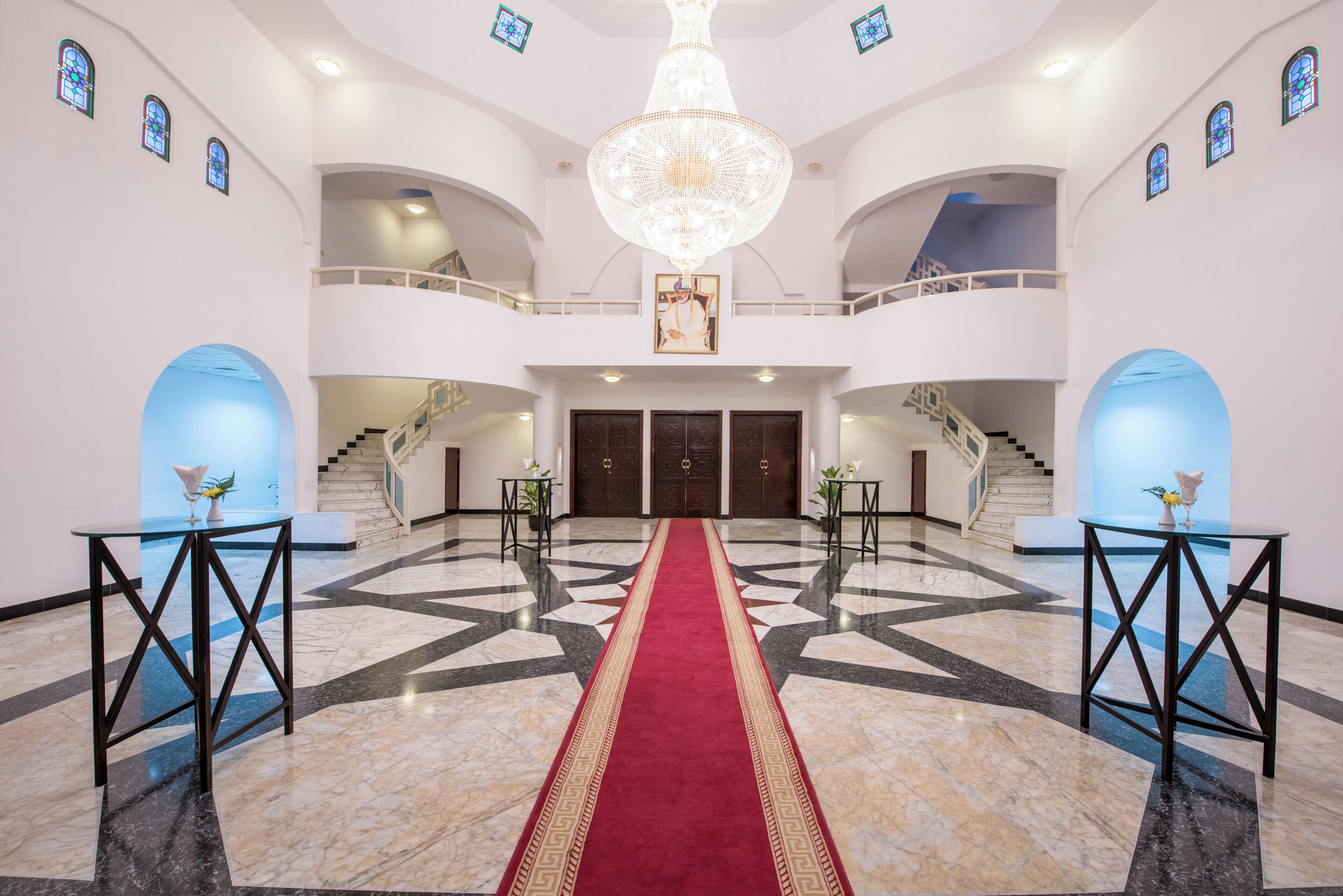 Un hall majestuoso con alfombra roja, pisos de mármol y un gran candelabro.