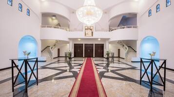 Un hall majestuoso con alfombra roja, pisos de mármol y un gran candelabro.