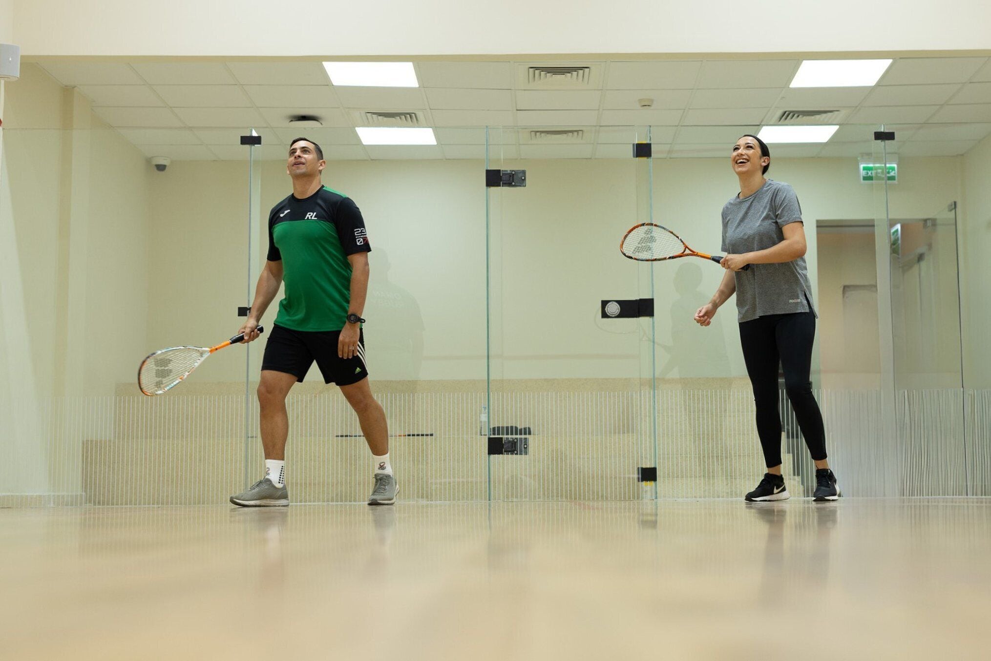 Due persone giocano a squash in una stanza con pareti in vetro e pavimento in legno.