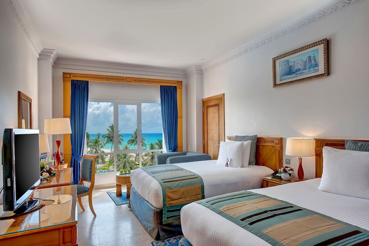 Habitación de hotel con dos camas, televisión, escritorio con lámpara y vista al océano.