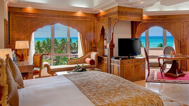 A luxurious hotel room with a large bed, a television, a desk with a chair, and a view of the ocean.