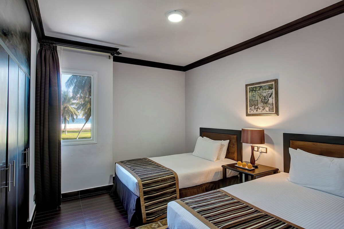 Habitación de hotel con dos camas, una ventana con vistas, un cuadro en la pared y una mesita de noche con una lámpara.