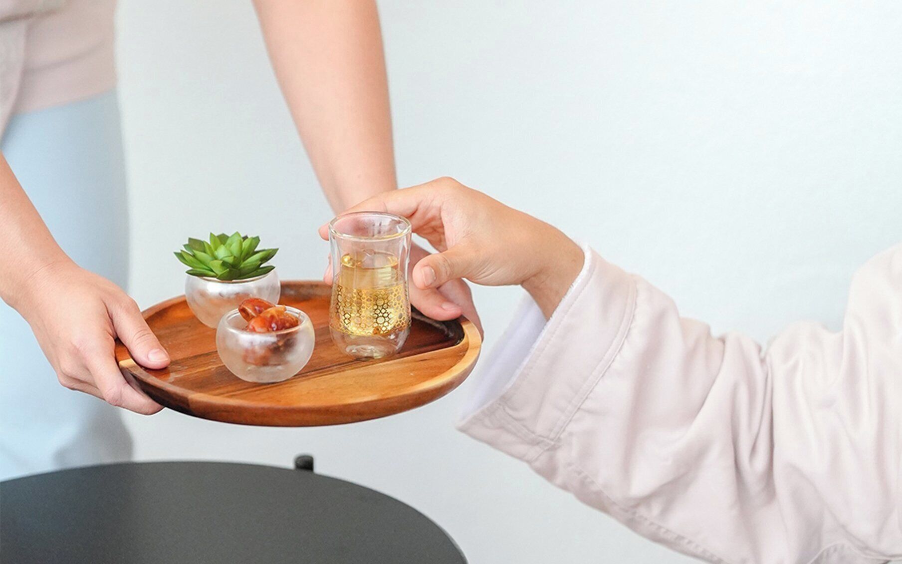 Una persona sosteniendo una bandeja con un vaso de líquido ámbar, una pequeña suculenta y un vaso transparente con un diseño dorado.