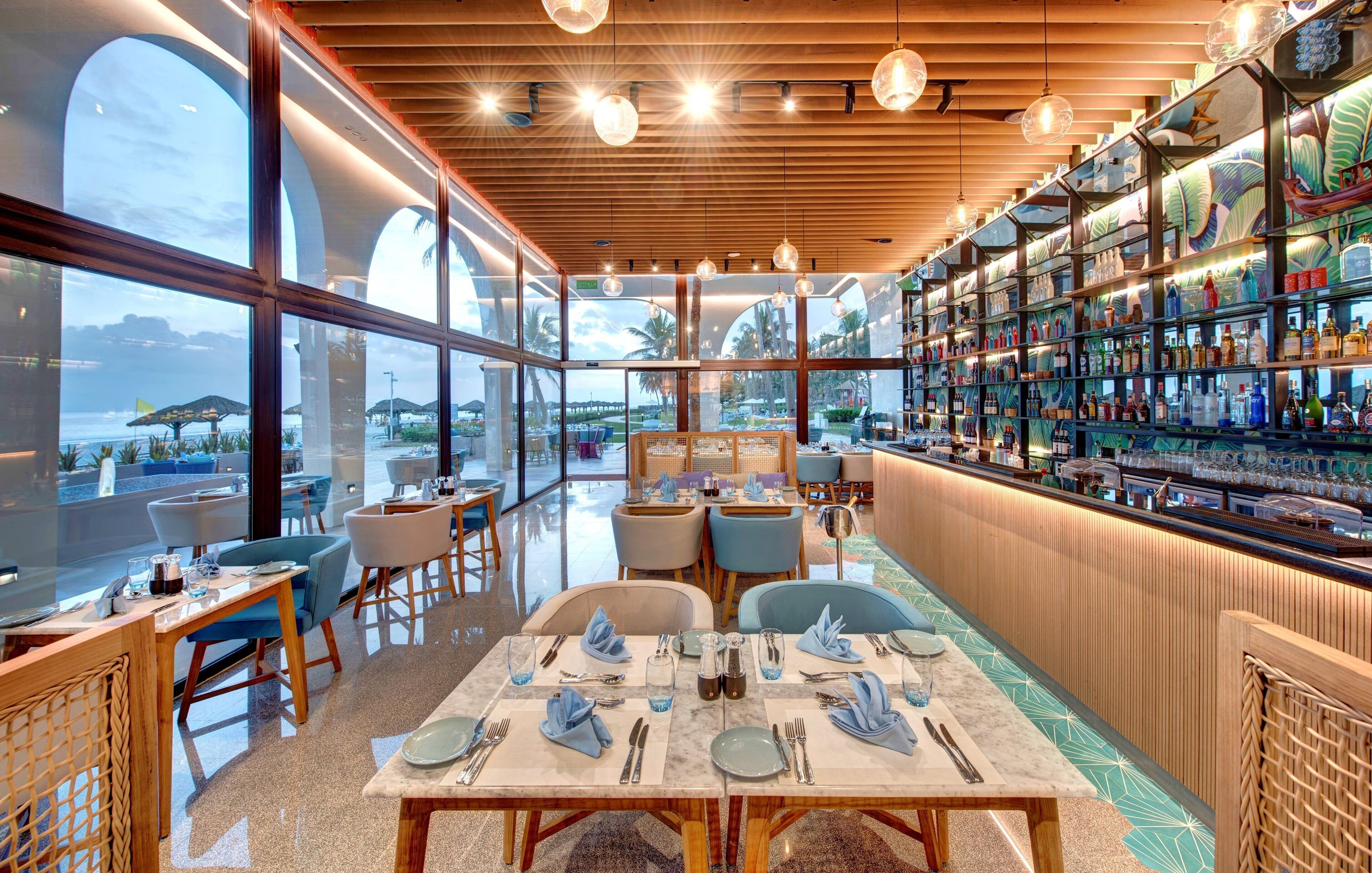 A restaurant with a view of the sea, wooden ceiling, and a bar with a variety of bottles.