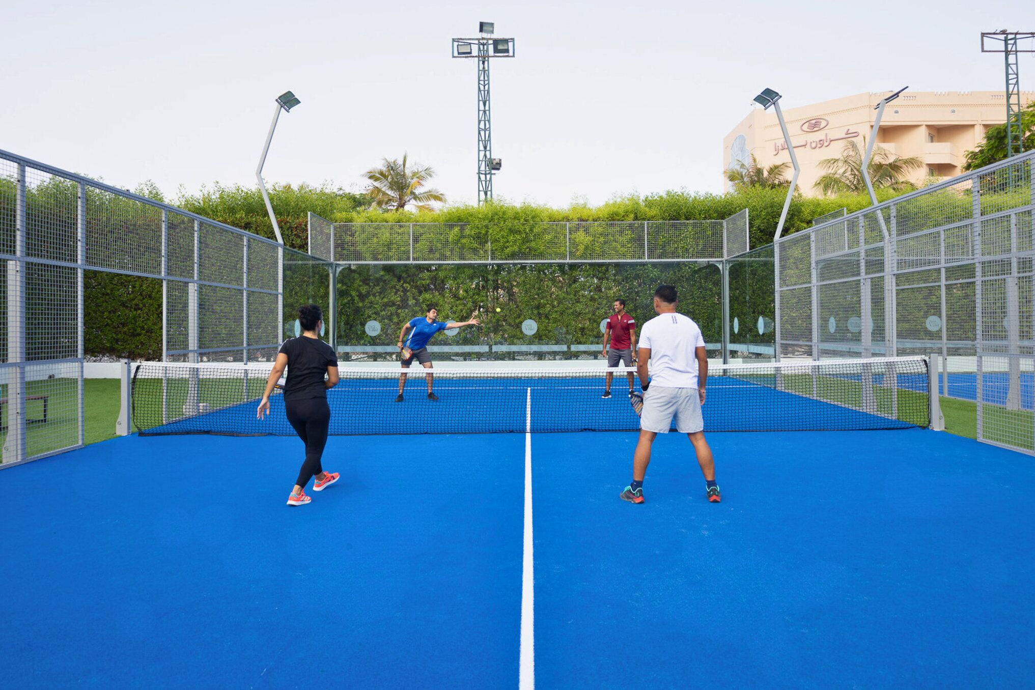 Una cancha de tenis con red, rodeada por una cerca metálica, y un edificio con escritura árabe al fondo.