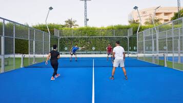 Una cancha de tenis con red, rodeada por una cerca metálica, y un edificio con escritura árabe al fondo.