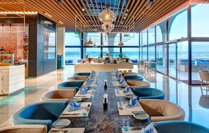 Un restaurant doté d’une longue table à manger, de chaises recouvertes d’un tissu bleu et offrant une vue sur l’océan grâce à de grandes fenêtres.