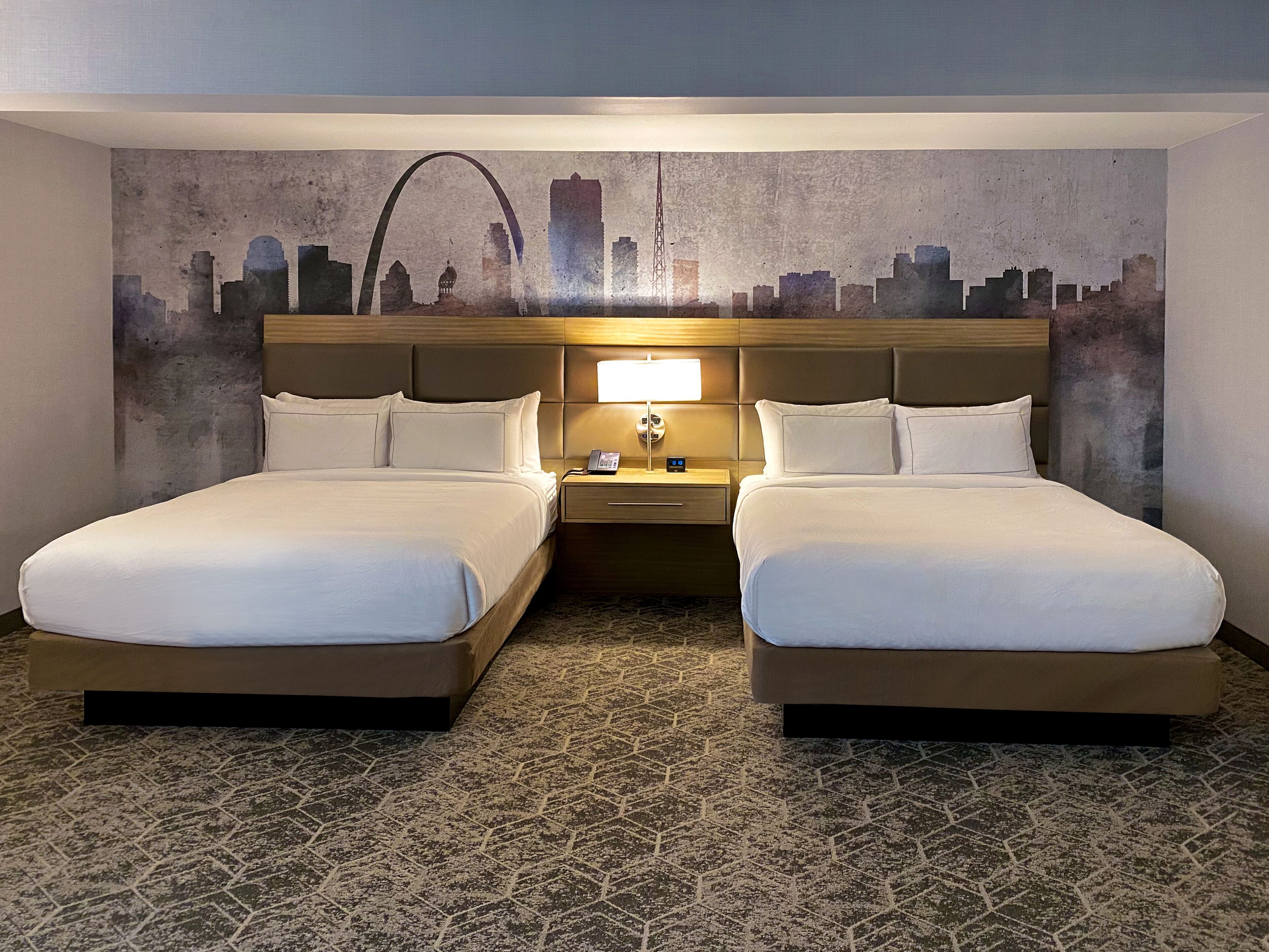 Ein Hotelzimmer mit zwei Betten, einem Nachttisch mit Lampe und einem Wandgemälde mit Stadtansicht.