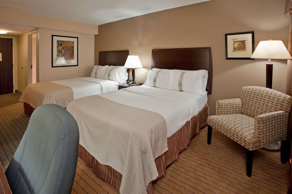 A hotel room with two beds, a chair, a desk, and a lamp.