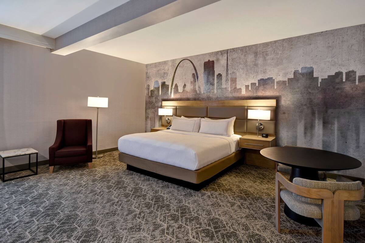 A hotel room with a large bed, a chair, a table, and a cityscape mural on the wall.