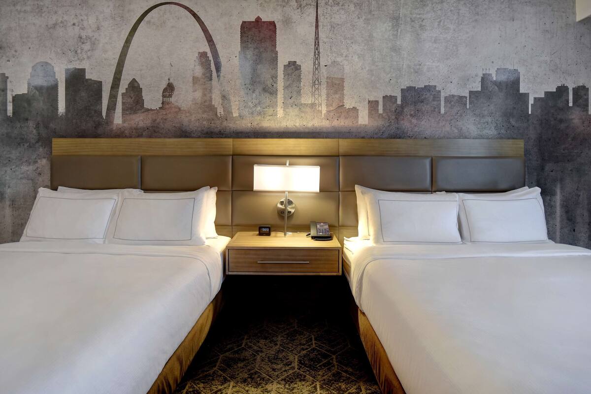 A hotel room with two beds, a city skyline mural on the wall, a wooden headboard, a nightstand with a lamp, and a patterned rug.