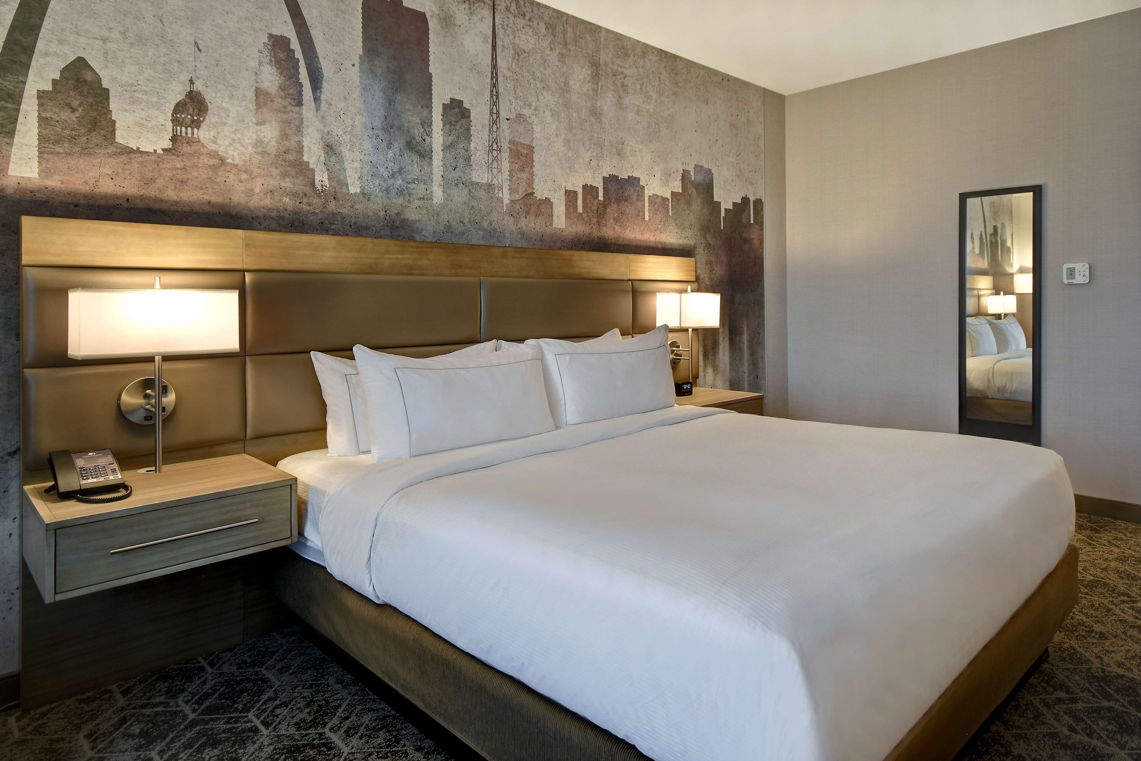 Habitación de hotel con una cama grande, dos mesitas de noche, un mural con un paisaje urbano en la pared y un espejo.