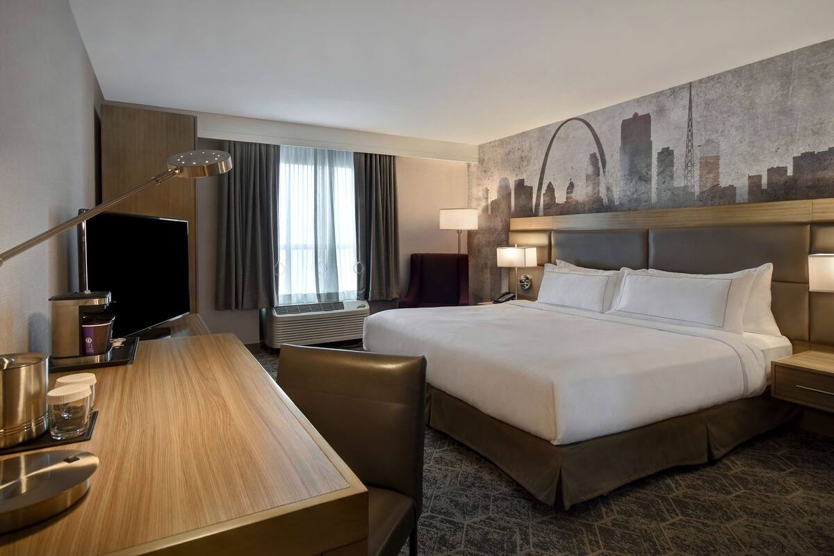 A hotel room with a large bed, a desk with a flat-screen TV, a coffee maker, and a cityscape mural on the wall.