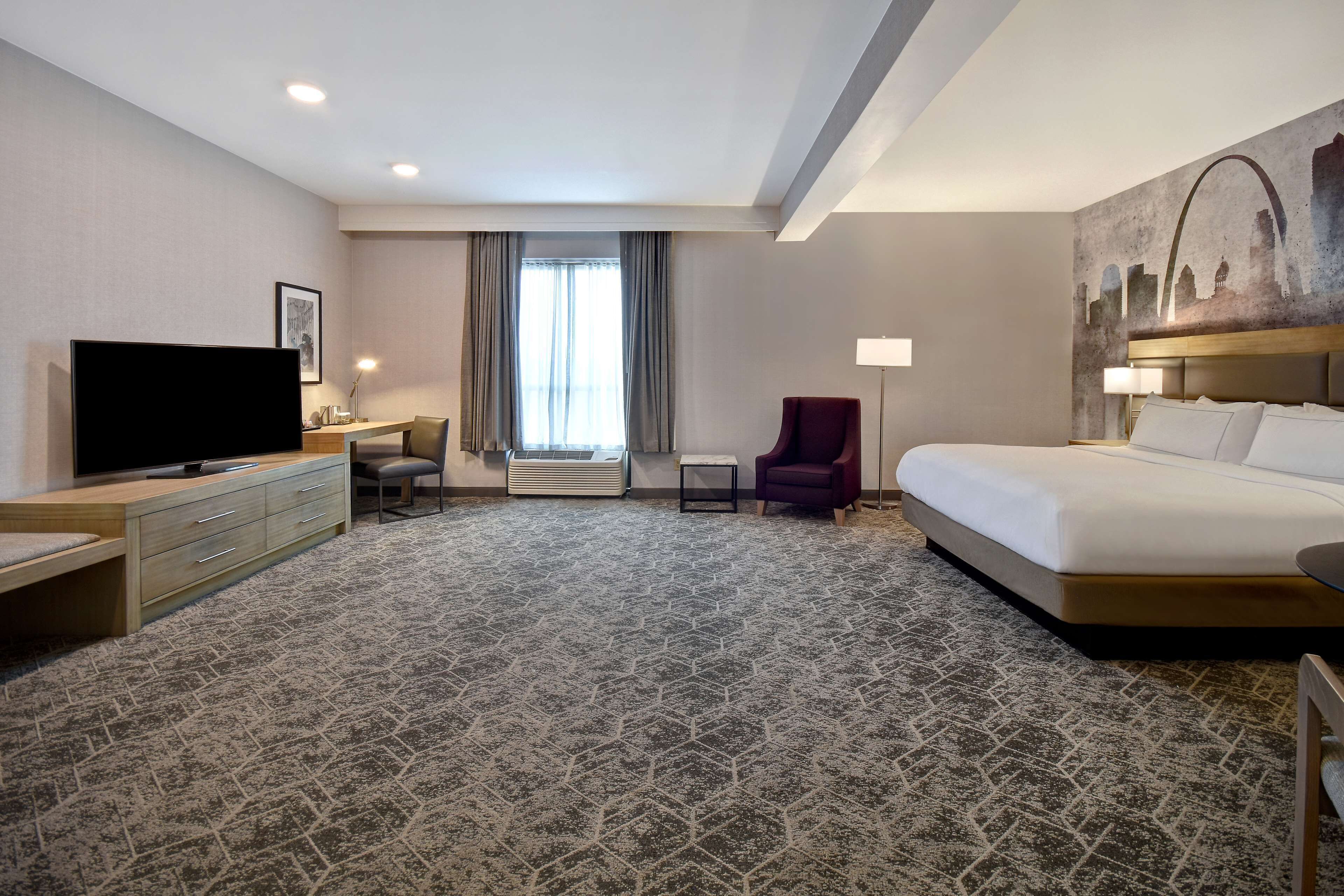 A hotel room with a large bed, a TV, a desk with a chair, a small table, and a window with curtains.