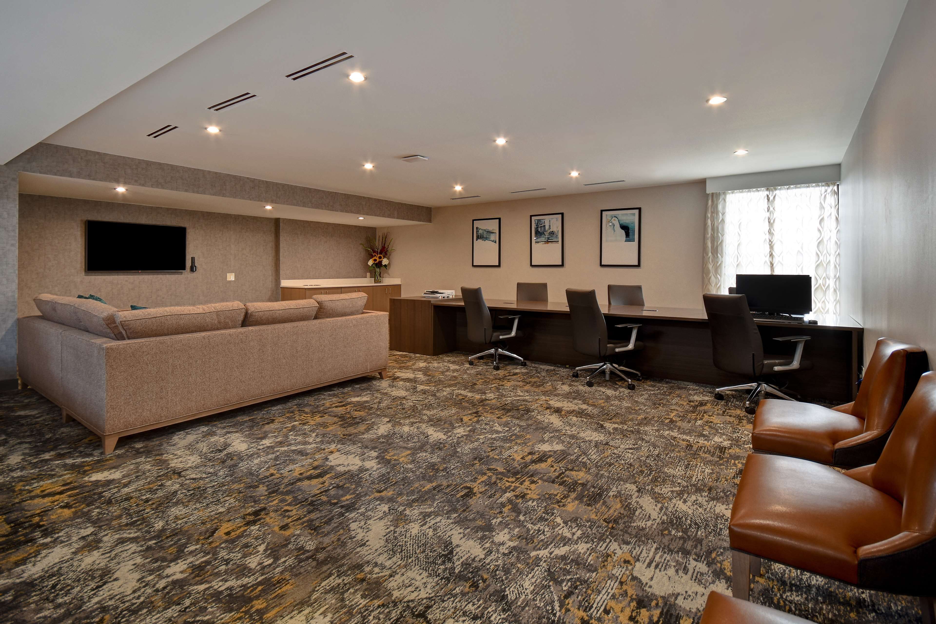 A spacious room with a large sofa, a dining table, and several chairs. There is a flat-screen TV mounted on the wall and a desk with a computer. The room has a patterned carpet and recessed ceiling lighting.