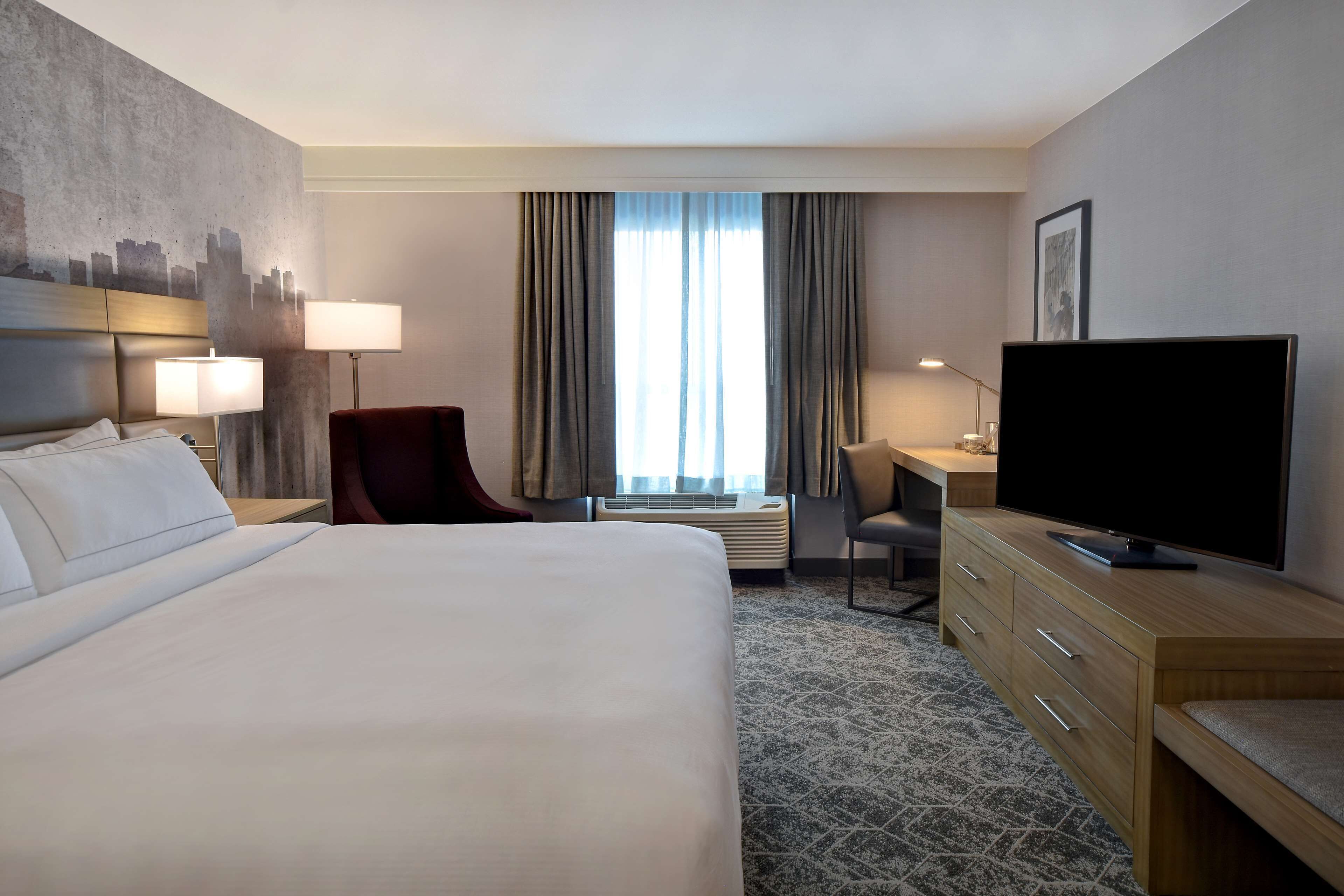 Ein Hotelzimmer mit einem großen Bett, einem Schreibtisch mit Stuhl, einem Fernseher und einem Stadtbild-Wandgemälde.