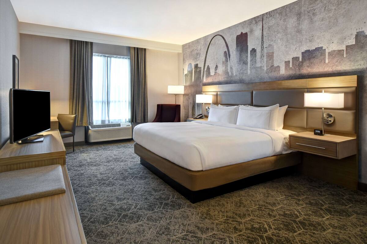 A hotel room with a large bed, a flat-screen TV, a desk, and a cityscape mural on the wall.