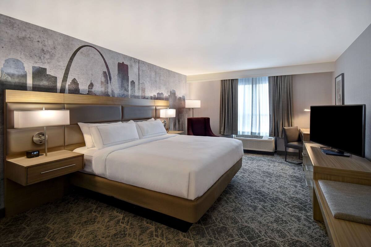 A hotel room with a large bed, a desk with a flat-screen TV, a chair, and a cityscape mural on the wall.