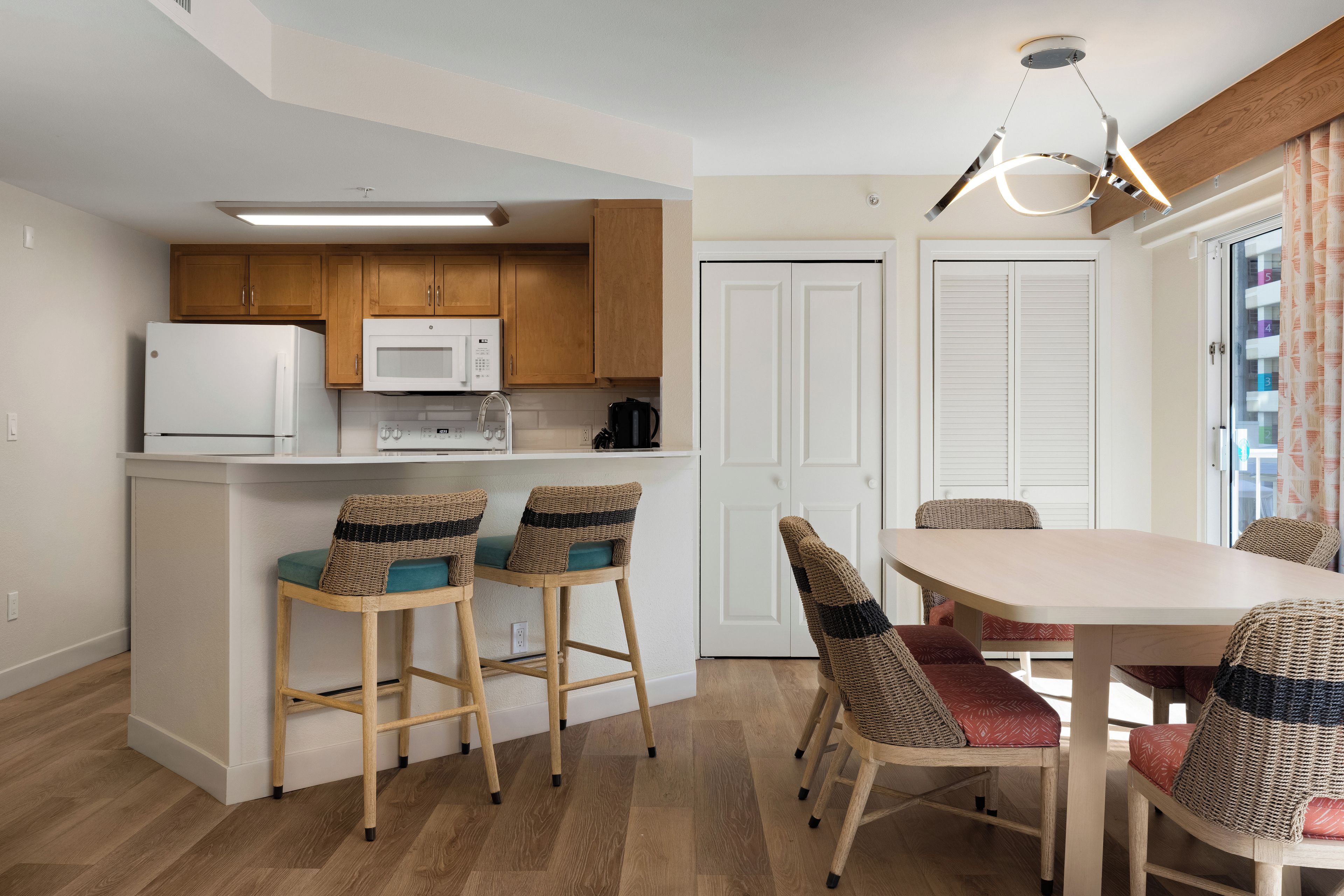 Cocina compacta con microondas, refrigerador y cafetera. Hay dos taburetes de bar y una mesa de comedor con sillas. El cuarto tiene piso de madera y paredes blancas.