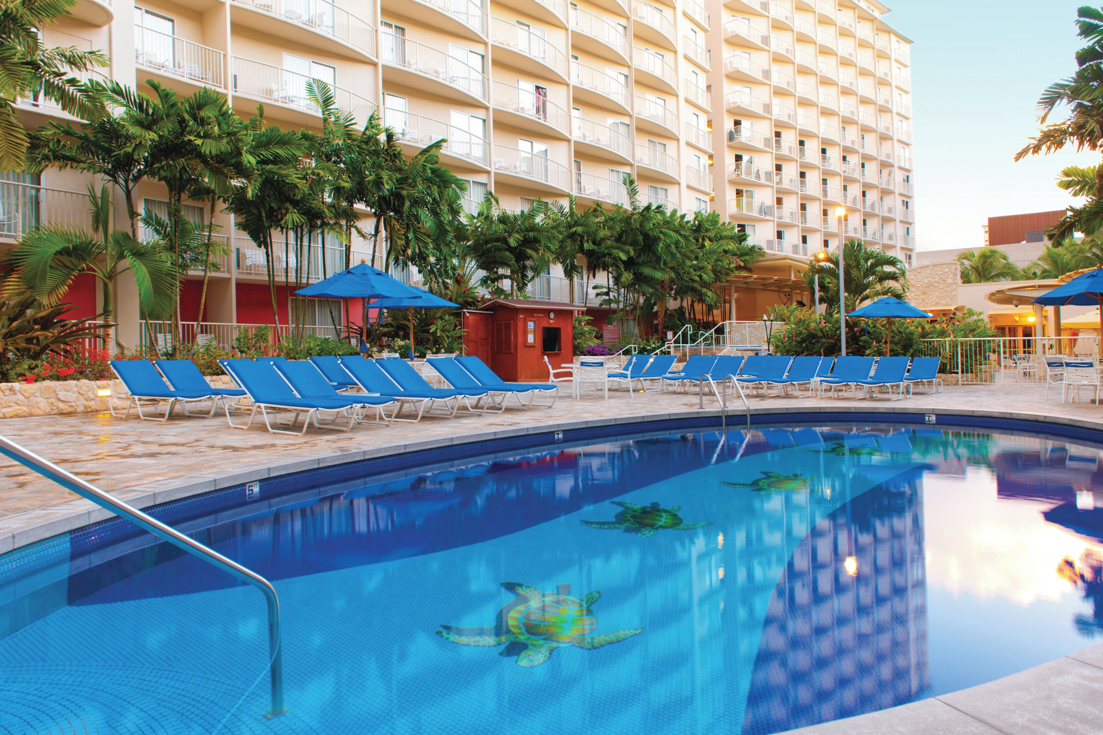 A hotel pool area with multiple lounge chairs and umbrellas, surrounded by palm trees and a multi-story building.
