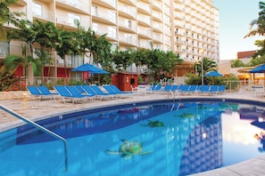 A hotel pool area with multiple lounge chairs and umbrellas, surrounded by palm trees and a multi-story building. - Club Wyndham at Waikiki Beach Walk (Honolulu)