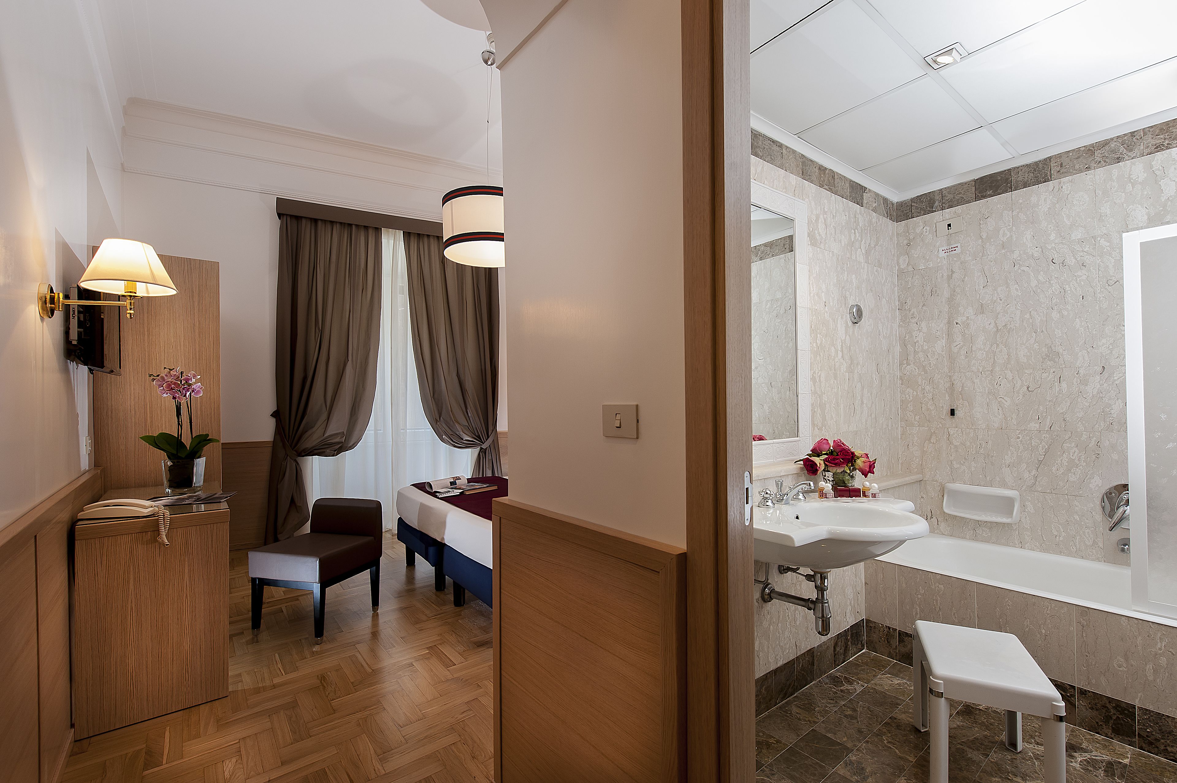 Une chambre d’hôtel avec un plancher en bois, un lit avec une tête de lit, une table de chevet avec une lampe, une petite armoire en bois et une fenêtre avec des rideaux.
