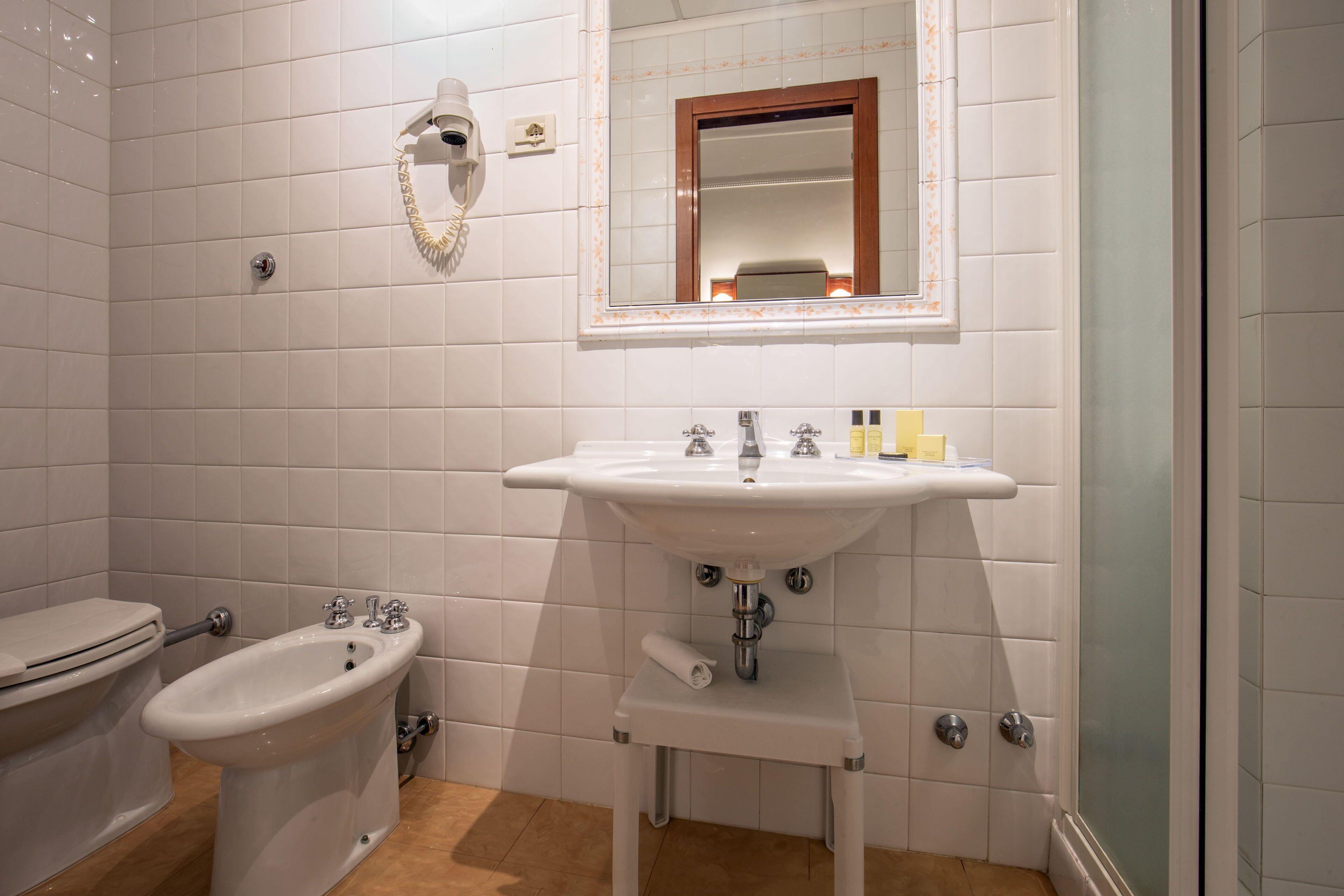 A bathroom with white tiles, a sink, a bidet, a toilet, and a mirror with a wooden frame.
