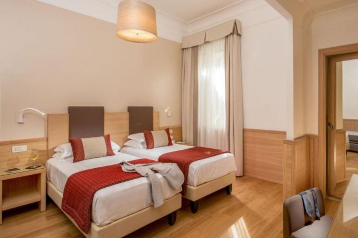 Een hotelkamer met twee bedden, een houten vloer, een bureau met lamp en een raam met gordijnen.