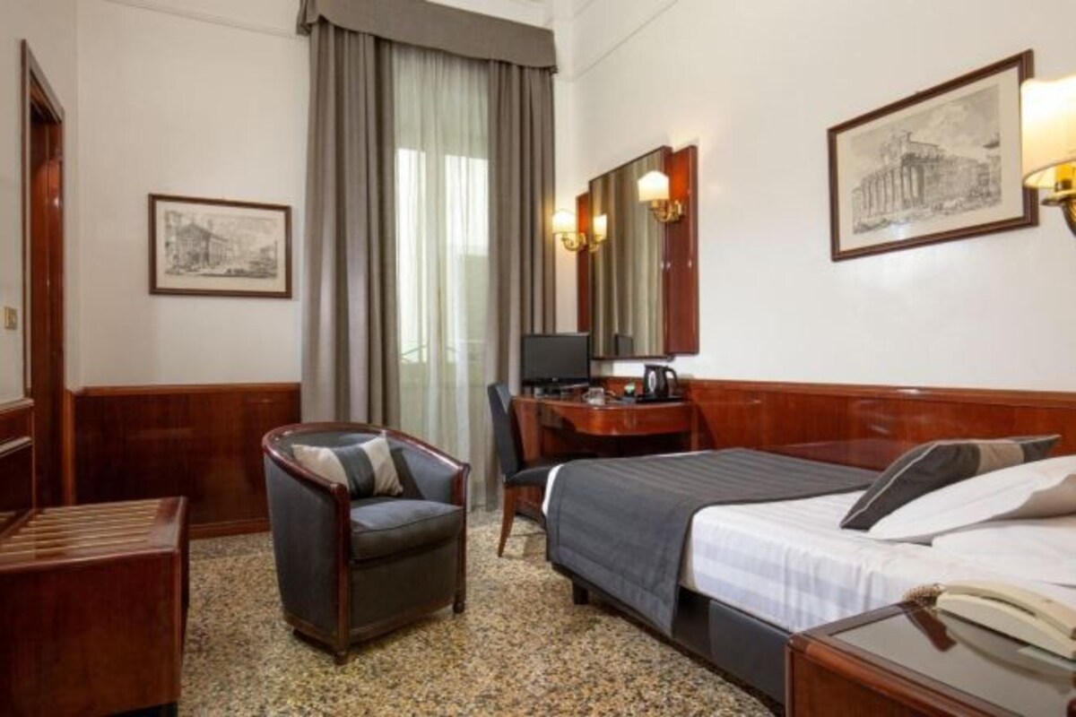 Een hotelkamer met een bed, een bureau met een computer, een stoel, een kleine tafel en een raam met gordijnen.