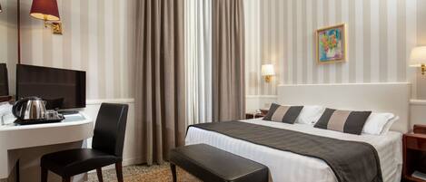A hotel room with a bed, desk, chair, and a small table with a lamp.
